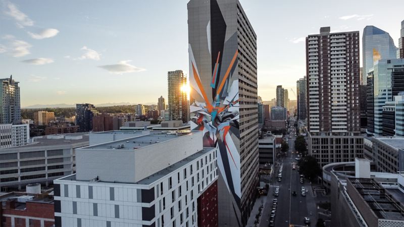 An aerial view of a cityscape at sunset with modern buildings, one featuring a large colorful mural, and the sun peeking from behind.