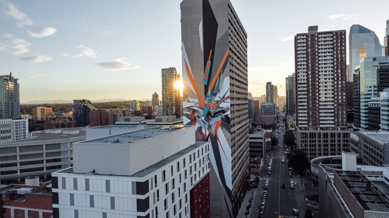 Vue aérienne du coucher de soleil sur une ville avec des immeubles modernes, dont un présente une grande murale colorée avec le soleil qui apparaît derrière.