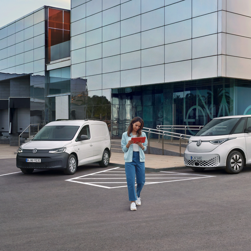 Frau mit Tablet in der Hand neben zwei weißen VW Nutzfahrzeugen, die vor einem modernen Gebäude parken