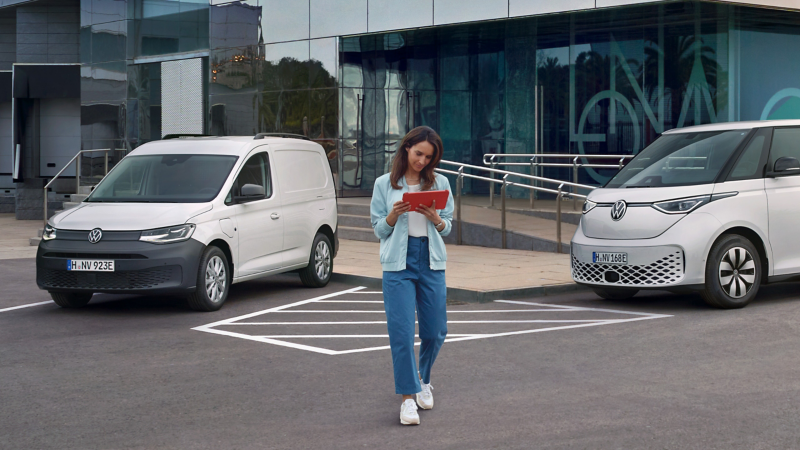 Eine Frau mit Tablet in der Hand neben zwei weißen VW Nutzfahrzeugen, die vor einem modernen Gebäude parken.