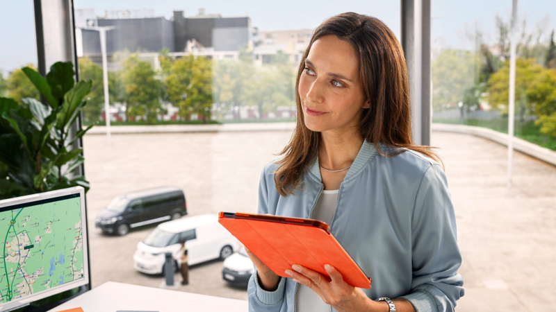 Eine Flottenmanagerin mit Tablet in der Hand im Büro, durch das Fenster sieht man VW Modelle auf dem Hof stehen.