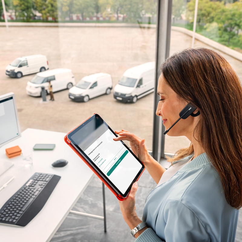 Eine Flottenmanagerin mit Headset sitzt am Computer und hält ein Tablet in der Hand. Im Hintergrund sieht man durch ein Fenster verschiedene VW Modelle.