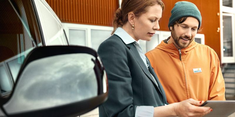 Zwei Personen stehen neben einem VW Transporter und blicken auf ein Tablet.
