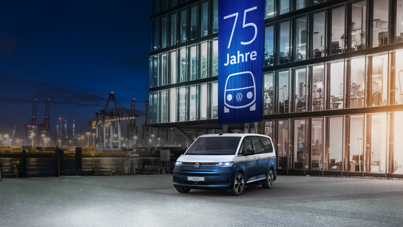 Ein blauer und weißer VW Multivan steht nachts vor einem beleuchteten Gebäude mit einem großen Banner zum "75 Jahre" Jubiläum.