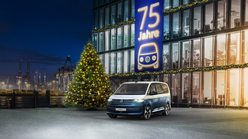 Ein blauer und weißer Volkswagen Multivan steht nachts vor einem festlich beleuchteten Gebäude mit einem großen Banner zum "75 Jahre" Jubiläum und einem Weihnachtsbaum davor.