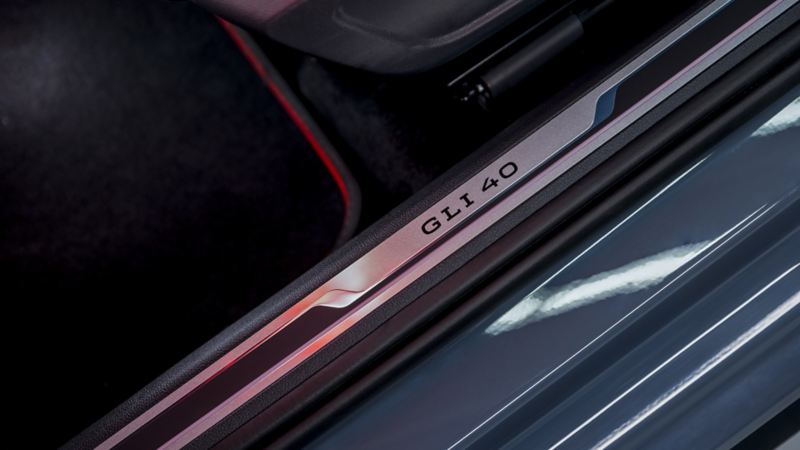 A Jetta GLI 2024 door sill. The door sill is black in color and has a metallic strip running along its length. The metallic strip has “GLI 40” written on it in black letters and the door sill is illuminated by a soft orange glow.