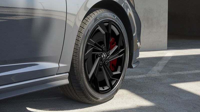 The Jetta GLI 2024 wheel in front of a concrete surface background. The wheel is black with a silver rim and a red brake caliper.
