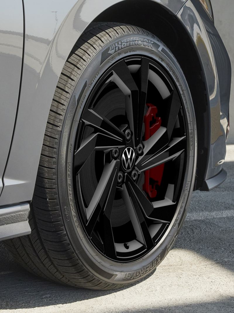 The Jetta GLI 2024 wheel in front of a concrete surface background. The wheel is black with a silver rim and a red brake caliper.