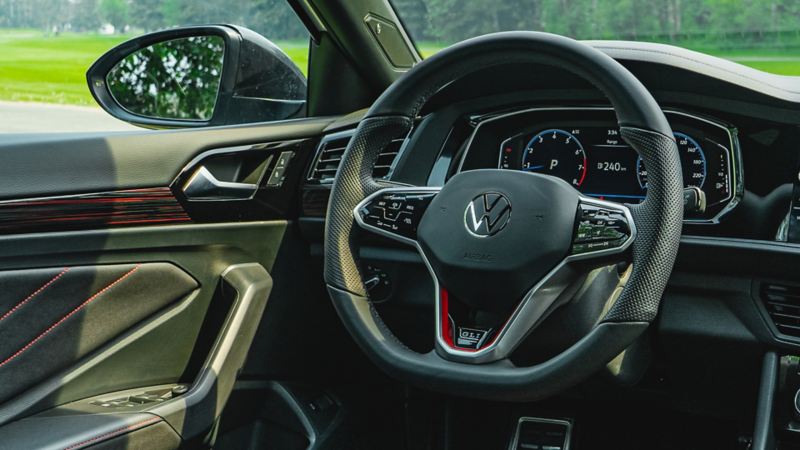 Jetta GLI 2024 black leather interior. The car is parked on a road with trees in the background. It has a large touchscreen display in the center console and a leather steering wheel.