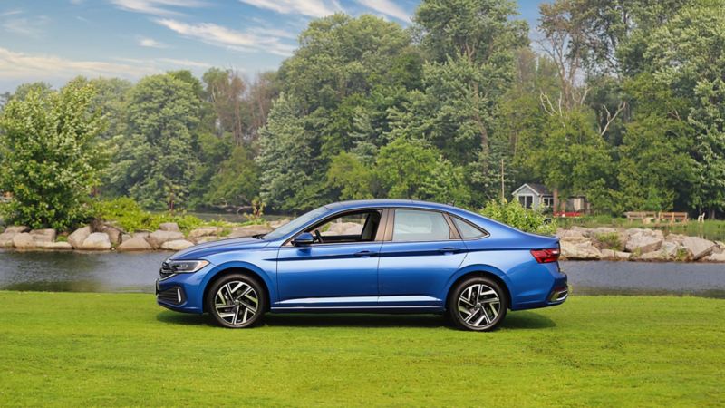 La Jetta 2024 bleue de Volkswagen garée sur une pelouse près d’un lac avec des arbres et des roches en arrière-plan sous un ciel bleu clair.