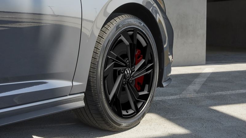Une roue de Jetta GLI 2024 devant un fond en béton avec l’ombre de la voiture. La roue est noire avec une jante argentée et un étrier de frein rouge. Le pneu est à profil bas.