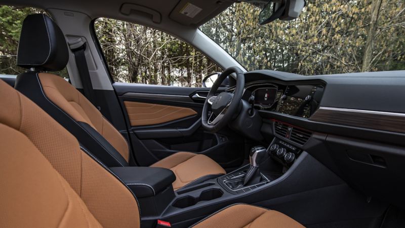 The interior of Volkswagen Jetta 2024 featuring black dashboard and a brown leather interior. The car has a sunroof and a touch screen display on the dashboard, gear shift in the center console and a cup holder, black steering wheel with buttons on it, black door panel with a silver handle.