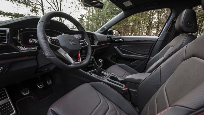 L’intérieur en cuir noir d’une Jetta GLI 2024. Le siège conducteur et le siège passager sont visibles sur l’image, ainsi que le tableau de bord et la console centrale. La voiture a un toit ouvrant et le volant porte le logo de Volkswagen. Les sièges ont des coutures rouges.
