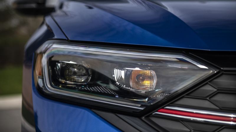 A close-up of the front headlight of Jetta GLI 2024. The headlight is rectangular in shape with a black border and has a clear lens with a yellow reflector on the side. The car has a black grille with a red stripe on it.