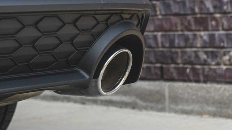 The rear end of a Jetta GLI 2024 with a brick wall in the background. The car is white in color and has a black bumper with a honeycomb pattern. The car has a chrome exhaust pipe.