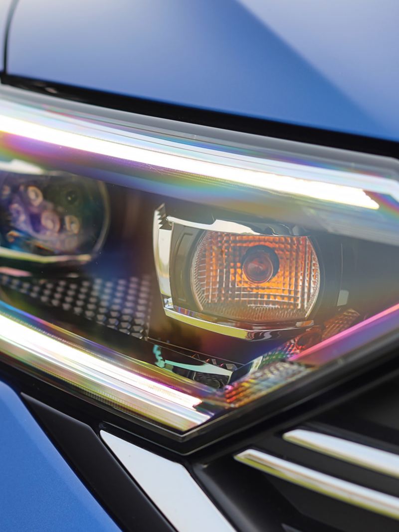 Close-up photo of the front headlight of a blue car Volkswagen Jetta 2024  with a modern design and a rainbow-like reflection on the surface. The headlight is turned on and the orange turn signal is visible