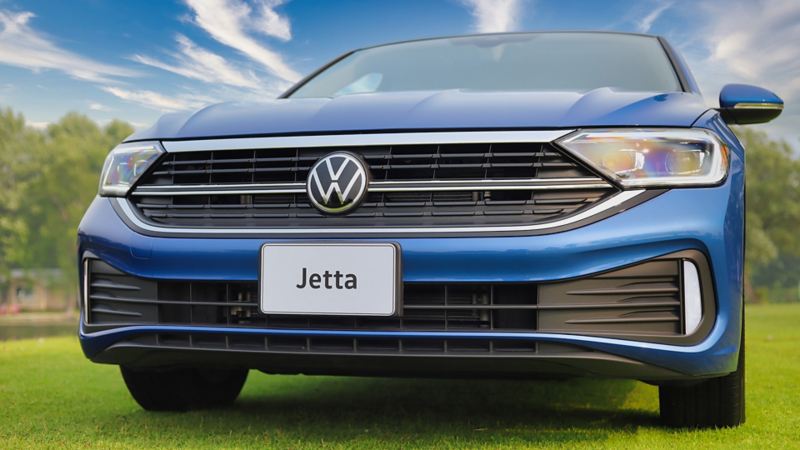 Blue Volkswagen Jetta 2024 car parked on a grassy field with a chrome grille and a Volkswagen emblem on the front. 