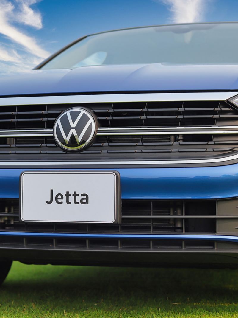 Blue Volkswagen Jetta 2024 car parked on a grassy field with a chrome grille and a Volkswagen emblem on the front. 