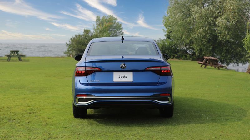 A rear view of the blue 2024 Volkswagen Jetta  parked on a grassy field near a lake with trees and picnic table in the background.