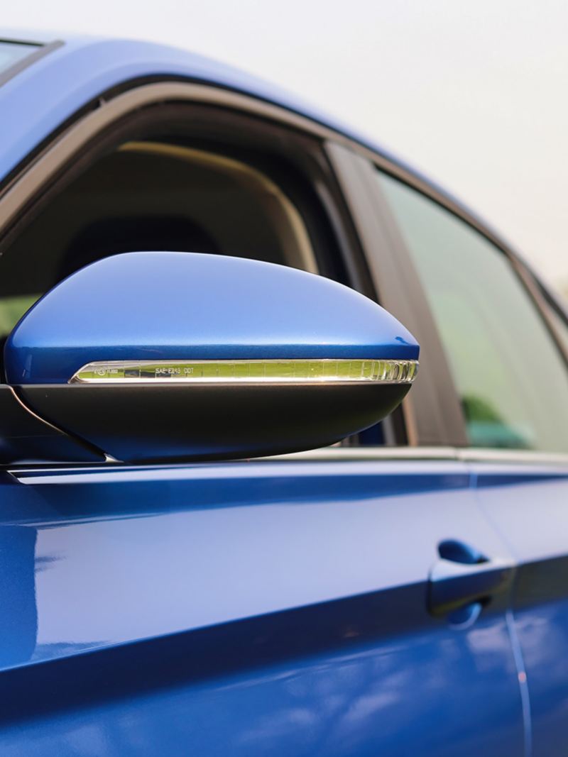 Volkswagen Jetta 2024 blue car’s side mirror and window. The side mirror is black and blue , has a silver trim. The window is rolled up and has a black tint.