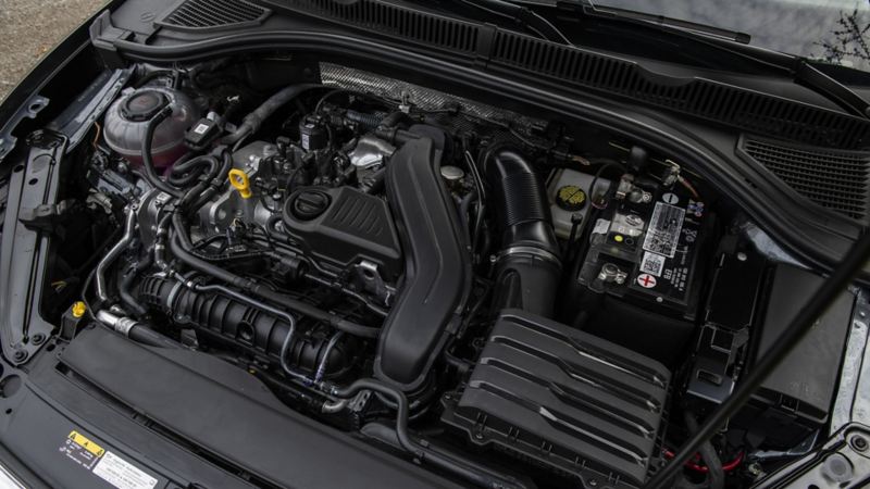 The photo of Volkswagen Jetta 2024 featuring the engine bay of a car. The engine is a V6 with a black plastic cover. The battery is located on the right side of the engine bay. The air filter is located on the left side of the engine bay. The car has a black radiator and a black plastic shroud, a yellow oil dipstick and a blue washer fluid cap. The hood is open and the hood prop is visible.