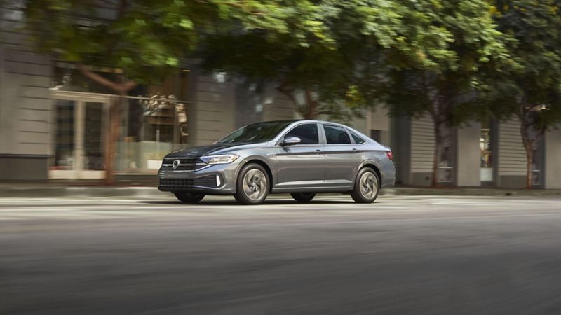 The photo of Volkswagen Jetta 2024 sedan driving on a city street. The car is driving on a black asphalt road with the background that consists of a modern building with a glass façade and trees lining the street.
