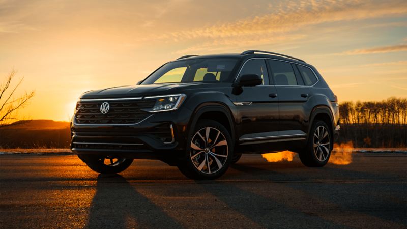 A Volkswagen Atlas 2025, parked on an open road with sparse trees in the background, showcases its design against a sunset sky with hues of orange and yellow.