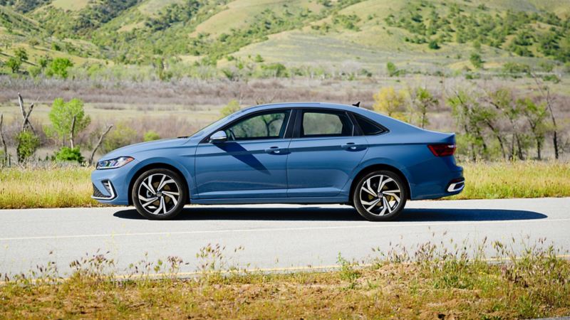 Vue latérale de la Volkswagen Jetta 2025 bleue stationnée sur une route pittoresque avec des collines et de la verdure en arrière-plan.