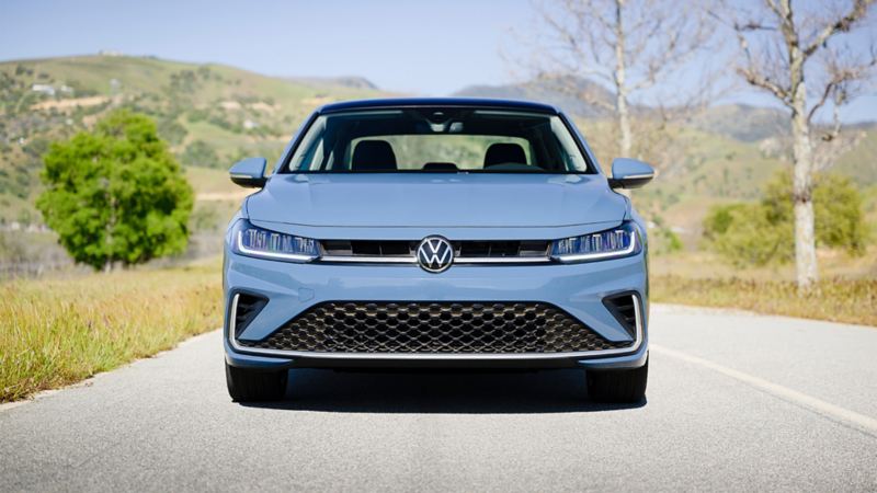 Front view of a blue VW Jetta 2025 parked on a scenic road with greenery on the background.