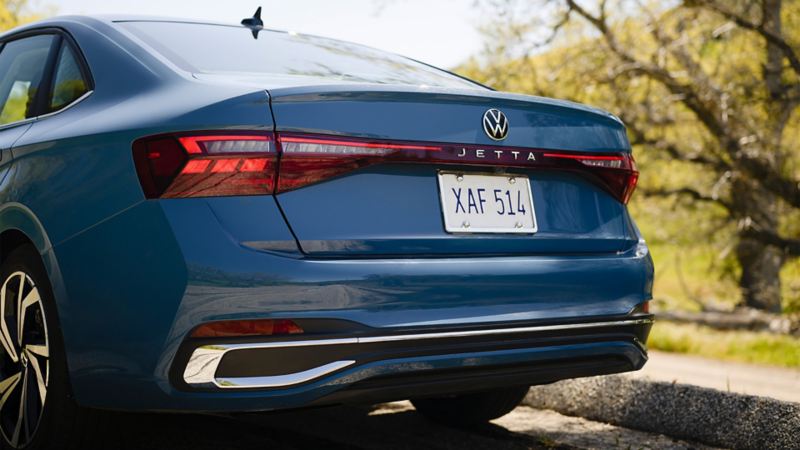 Rear view of a blue Jetta 2025 parked outdoors with a greenery in the background.