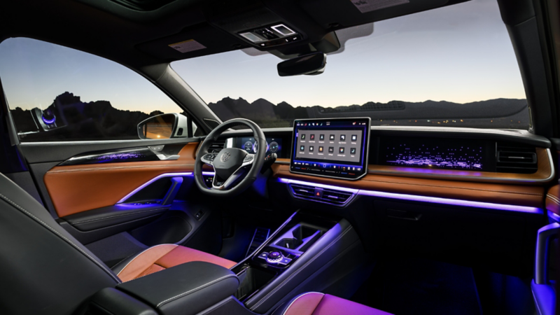 The interior of the all-new 2025 Tiguan, displaying the front seats, steering wheel, dashboard and infotainment screen, illuminated with purple interior ambient lighting.