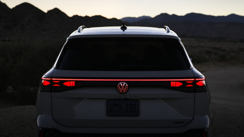 A back view of the white all-new 2025 Tiguan. It is dark so the vehicle and background isn’t very visible, and the rear taillight and VW logo are illuminated in red. 