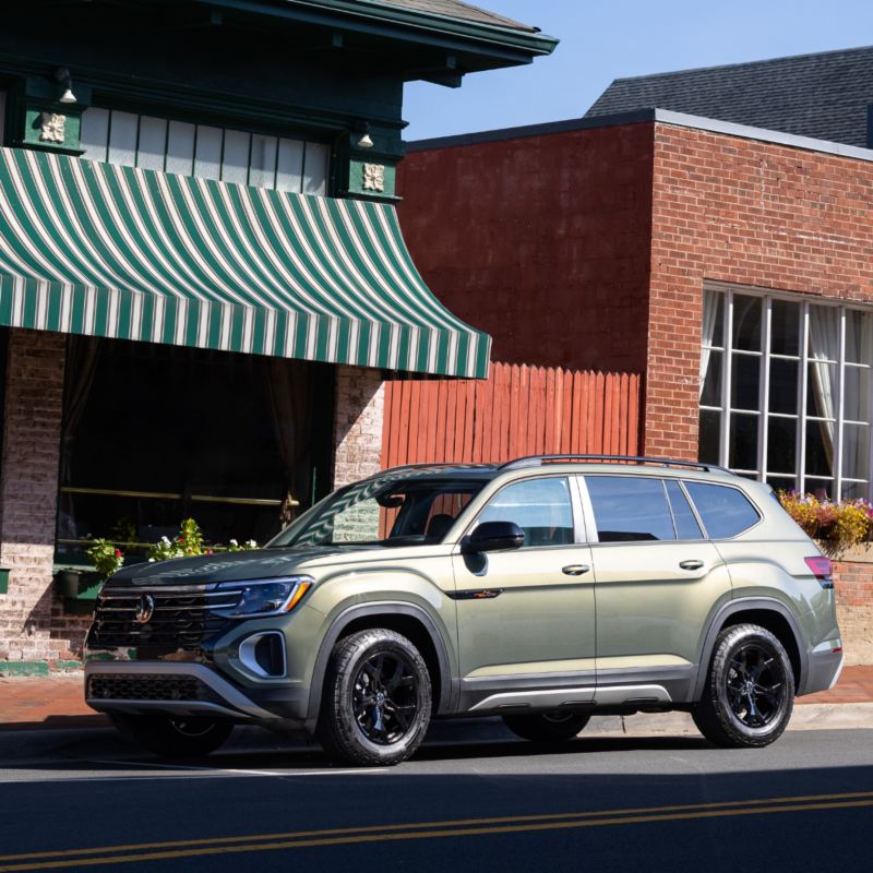 A shiny Avocado Green Pearl SUV is parked on a street beside a sidewalk, with buildings featuring brick walls and green awnings in the background.