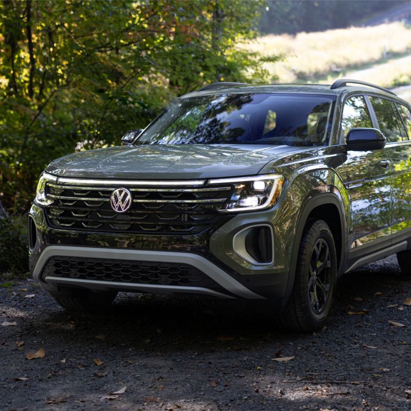 A Volkswagen Atlas 2025 Peak Edition, showcasing its aggressive design with a redesigned front grille, and bold wheel arch trims is parked on a gravel path surrounded by lush greenery.