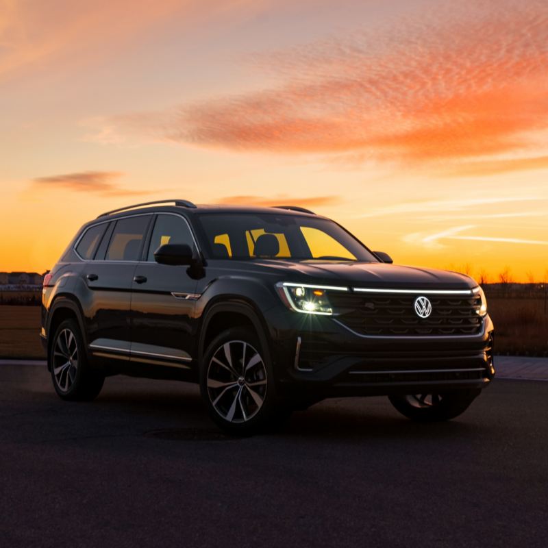 A Volkswagen Atlas 2025, bathed in the warm colours of a setting sun, showcases its design and headlights against an orange and pink sky.