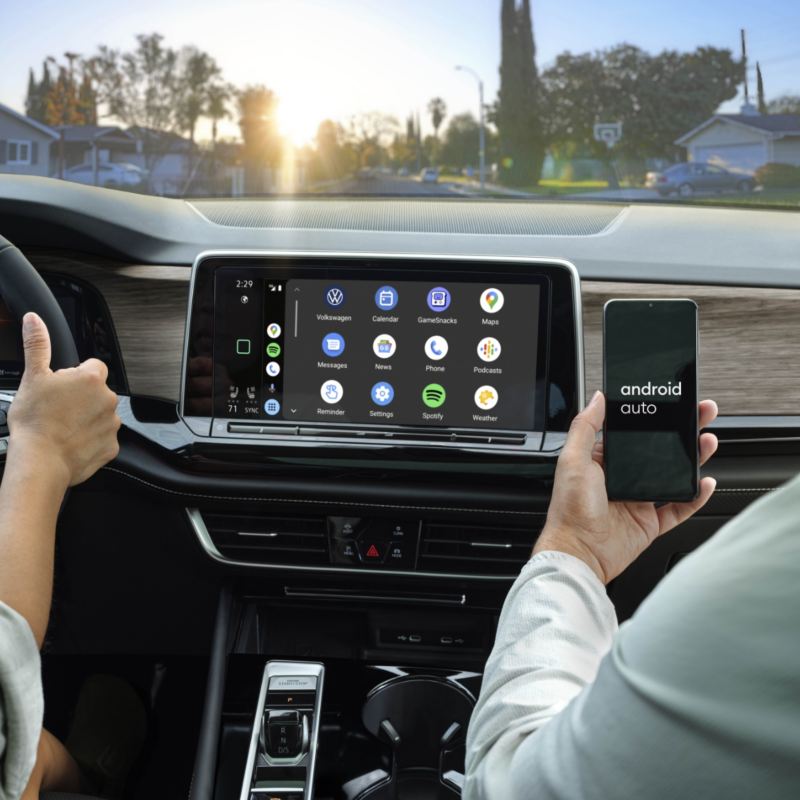 A passenger's hand holds a smartphone displaying "Android Auto," connected to the VW Atlas 2025's infotainment system, showcasing a variety of app icons on the touchscreen.