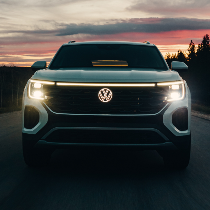 A front view of 2025 Atlas Cross Sport with its headlights illuminated, stands out against a vibrant twilight sky painted with hues of pink and orange.
