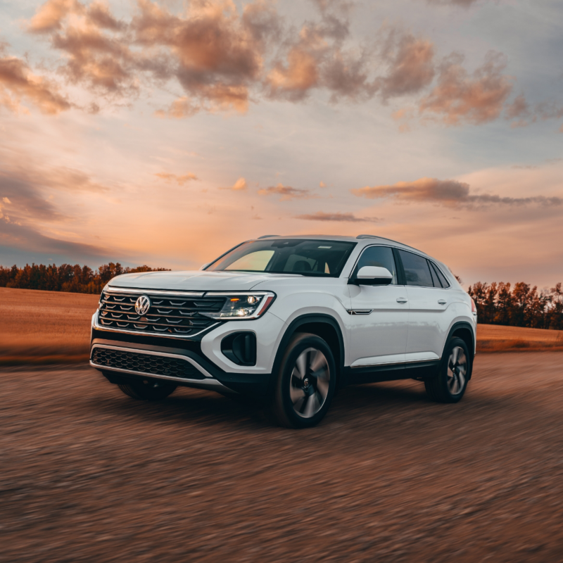 A white 2025 Atlas Cross Sport drives on a road with a sunset sky in the background, featuring hues of orange and blue.