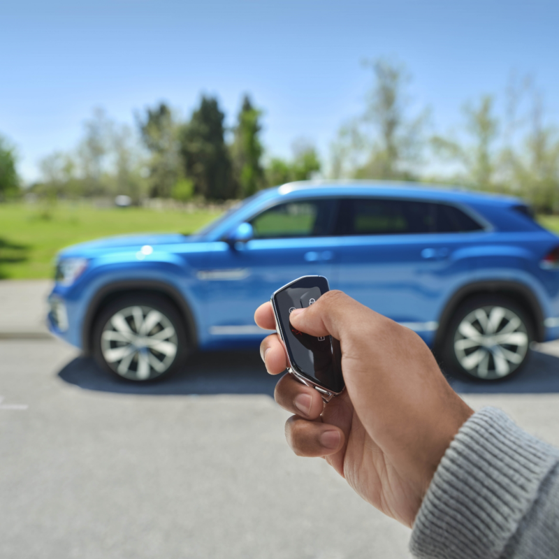 A hand holds a car key fob, ready to unlock a blue 2025 Atlas Cross Sport seen in the background.
