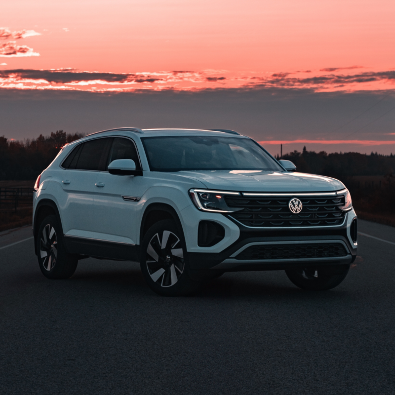 A white Atlas Cross Sport 2025 is parked on a road with its headlights on, with a pink and orange sunset sky in the background.