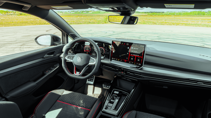 Vue côté conducteur de l’intérieur de la Volkswagen Golf GTI 2026, avec le volant, le tableau de bord numérique, l’écran tactile et les sièges sport noirs avec accents rouges.