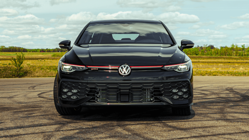 Front view of a black 2026 Volkswagen Golf GTI parked on pavement, featuring LED headlights and a red grille accent. 