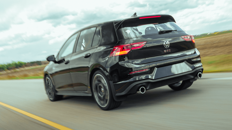 Rear view of black 2026 Volkswagen Golf GTI driving on open road, showing dual exhausts, GTI badge, and sporty rear design. 