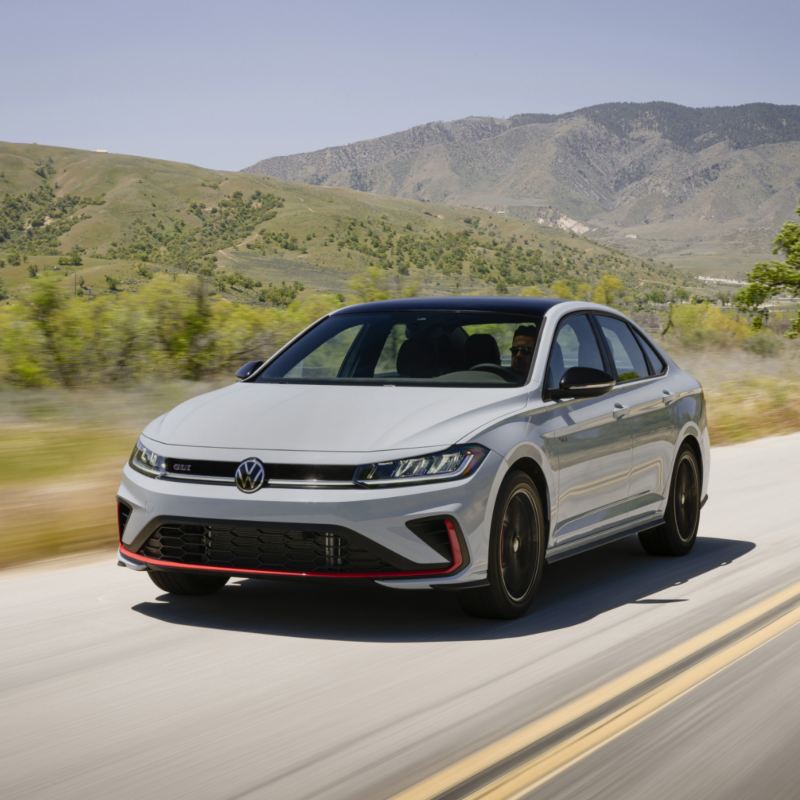 A white Volkswagen Jetta GLI 2025drives along a scenic road, with lush green hills in the background.