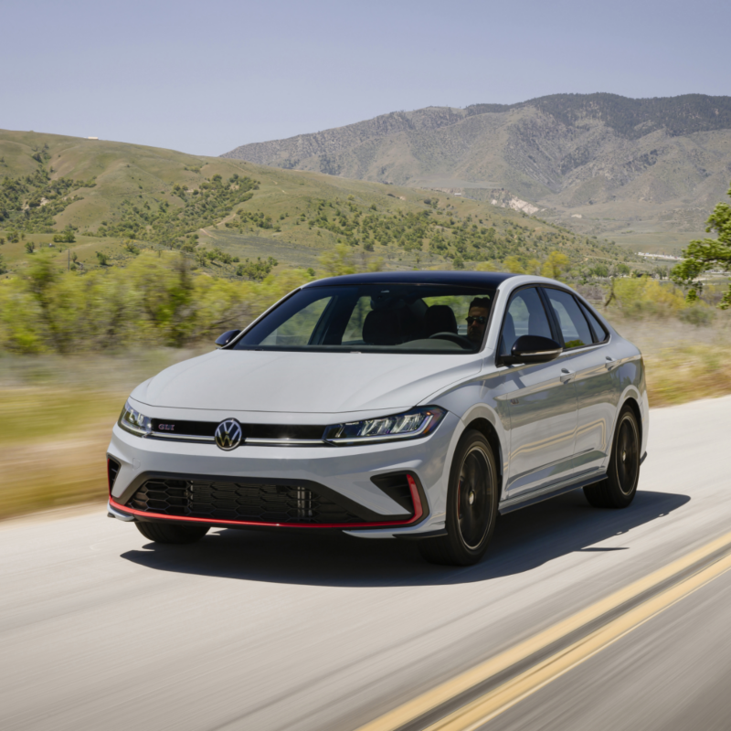 A white Volkswagen Jetta GLI 2025 drives along a scenic road, with lush green hills in the background.