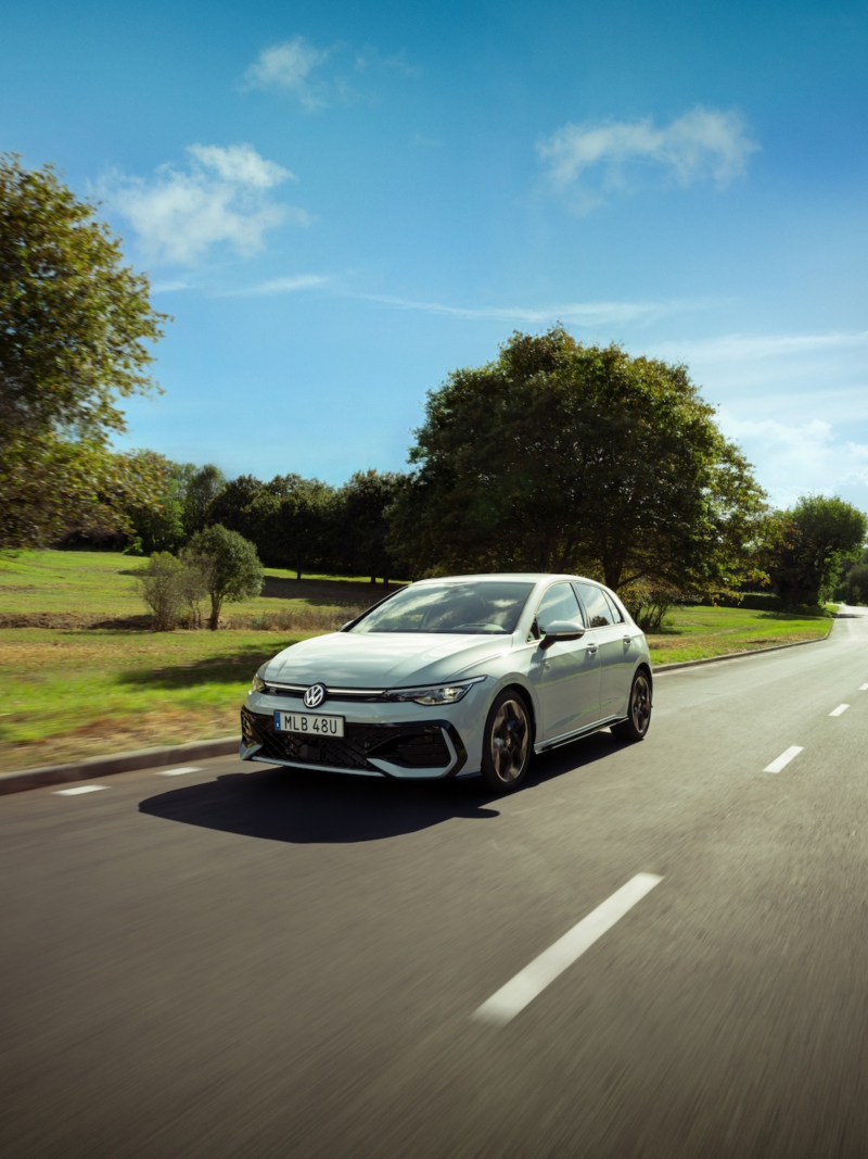 En vit VW Golf kör längs en väg med träd i bakgrunden.