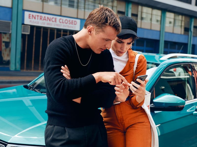 Paar lehnt an einen blauen VW T-Cross und schauen gemeinsam auf ein Smartphone
