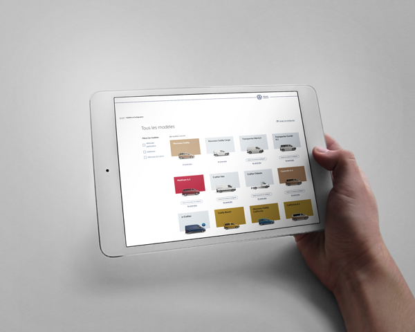 Une main tient une tablette affichant la page d'un configurateur de véhicules Volkswagen Utilitaires, permettant de choisir et de personnaliser différents modèles de voitures.