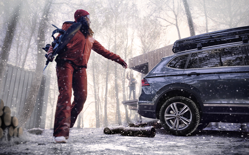 Une femme en tenue de sports d'hiver porte un snowboard et marche à côté de son véhicule Volkswagen, sous la neige