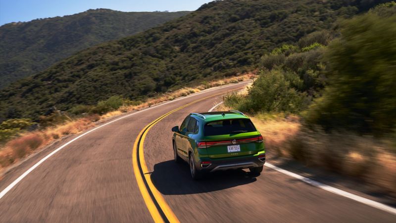 A wide shot of the rear of a Serrano Green 2025 Taos, driving on a hilly highway.